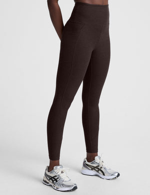 woman wearing brown high-waisted midi length leggings with pockets on a plain background. 