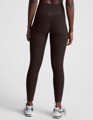 woman wearing brown high-waisted midi length leggings with pockets on a plain background. 