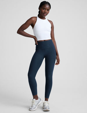 woman wearing a white high-neck racerback cropped tank and navy leggings on a plain background. 