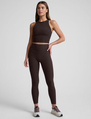 woman wearing a brown high neck racerback cropped tank and leggings on a plain background. 