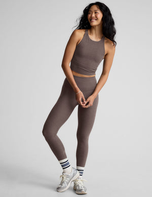 Woman wearing a brown high-neck racerback cropped tank and leggings against a gray background. 
