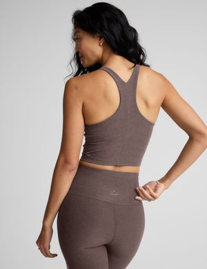 Woman wearing a brown high-neck racerback cropped tank and leggings against a gray background. 