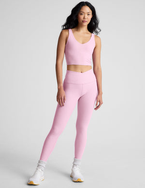 Woman wearing a pink v-neck cropped tank and leggings against a plain background. 