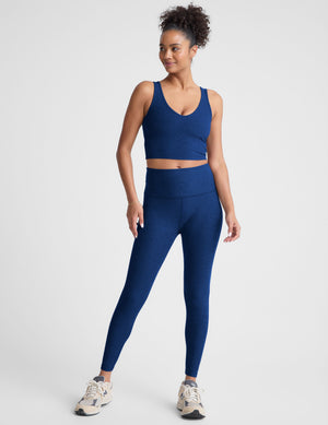 Woman wearing a blue v-neck cropped tank and leggings on a light gray background. 