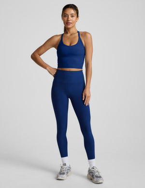 Woman wearing a blue high racerback cropped tank and leggings on a plain background. 