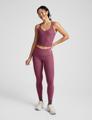Woman wearing a purple v-neck tank top with crossover back straps and leggings against a plain background. 