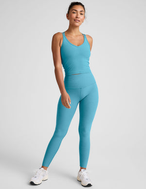 Woman wearing a blue tank top with crossover straps on back and leggings against a light gray background. 