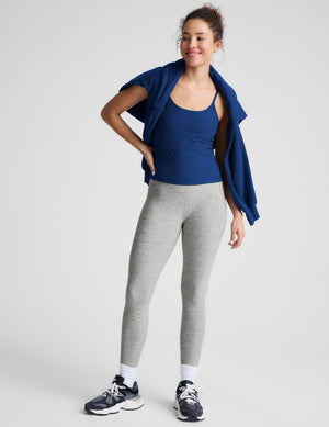 Woman wearing a blue tank top and gray leggings against a light gray background. 