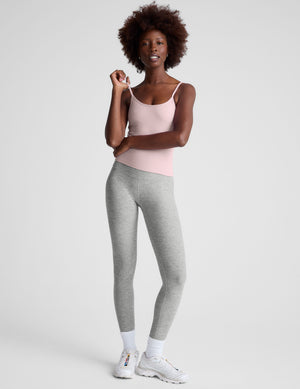 Woman wearing a light pink tank top and gray leggings against a plain background. 