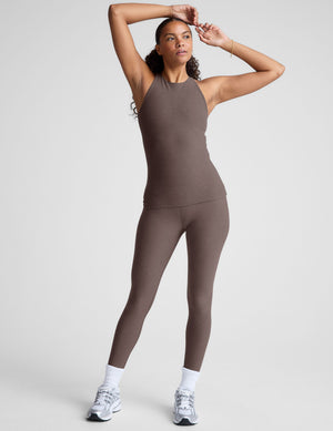 Woman wearing a brown high neck racerback tank top and leggings on a light gray background. 