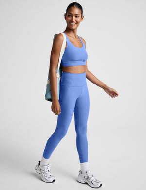 Woman wearing a blue cropped tank with a crossover back detail and blue leggings against a plain background. 