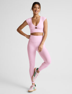 Woman in pink cropped twist tee and leggings on a light gray background. 