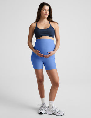 Woman wearing a blue bra and maternity shorts against a plain background. 