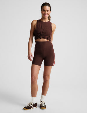 Woman wearing a brown tank top and brown biker shorts with pockets against a plain background. 