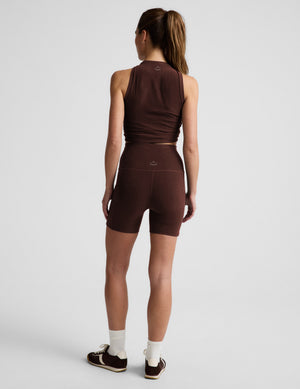 Woman wearing a brown tank top and brown biker shorts with pockets against a plain background. 