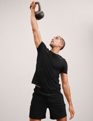 man lifting a kettlebell above his head wearing a black t-shirt and black spacedye shorts against a plain background. 