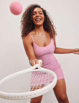 model is wearing a pink mini length dress with slim adjustable straps and built-in shorts with pockets. 