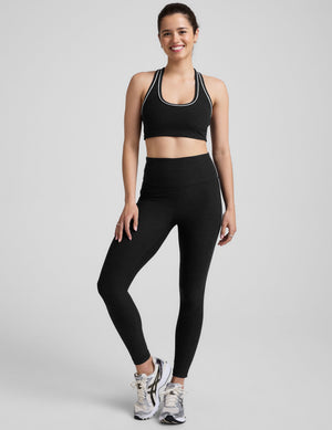 model wearing a black scoop neck racerback sports bra and leggings with white trim on a gray background. 