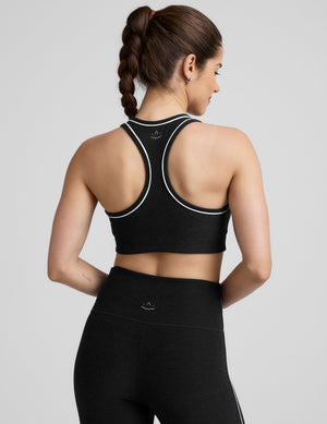 model wearing a black scoop neck racerback sports bra and leggings with white trim on a gray background. 