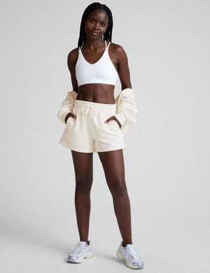 woman wearing a white bra with back crossover straps and white fleece shorts against a plain background. 