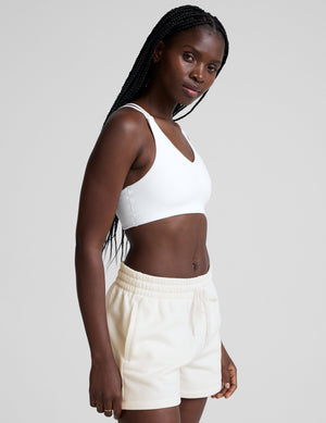 woman wearing a white bra with back crossover straps and white fleece shorts against a plain background. 