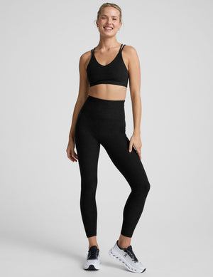 woman wearing a black sports bra with back crossover straps and leggings on a plain background. 