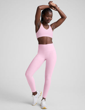 Woman wearing a pink v-neck bra with crossover back straps and leggings on a plain background. 