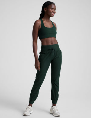 model wearing a green scoop neck racerback long line sports bra and joggers against a plain background. 