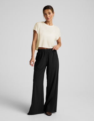 Woman wearing a cream colored cropped tee and black pants against a plain background. 