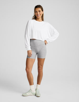 Woman wearing a white long sleeve and gray shorts on a plain background. 