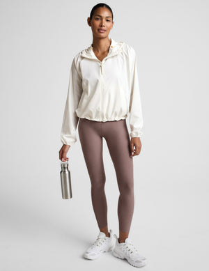 Woman wearing a white jacket and brown leggings, holding a water bottle on a light gray background. 