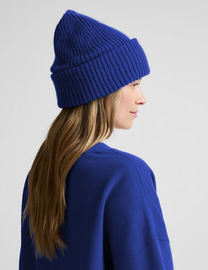 A woman shown from the side and back wearing a vibrant royal blue ribbed knit beanie and a matching royal blue sweatshirt, set against a plain light gray studio background.
