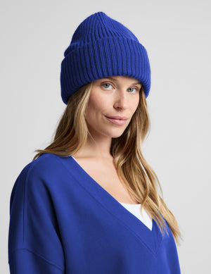 A woman wearing a vibrant royal blue ribbed knit beanie and a matching blue V-neck sweater, photographed against a neutral light gray background.