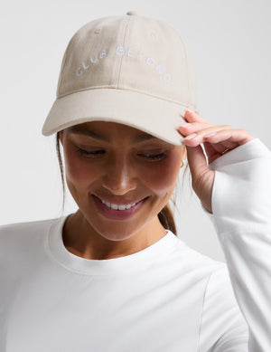 A woman wearing a beige baseball cap with 'CLUB BEYOND' embroidered in white on the front, paired with a white long-sleeve top against a light gray background.