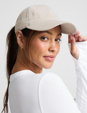 A woman wearing a beige baseball cap with 'CLUB BEYOND' embroidered in white on the front, paired with a white long-sleeve top against a light gray background.