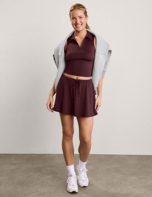 A woman modeling a dark burgundy athletic set consisting of a sleeveless collared crop top and a matching mini skirt, styled with a light gray sweatshirt draped over her shoulders, white socks, and white sneakers against a neutral studio backdrop.