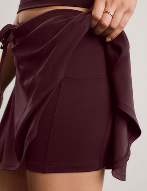Close-up of a person wearing a dark burgundy athletic mini skirt with a pocket, holding the fabric up to show built-in shorts. A matching top is partially visible at the top edge.
