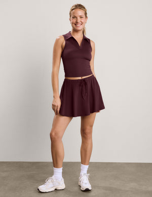A woman modeling a sleeveless collared athletic top and matching flared mini skirt in a deep plum-burgundy color, paired with white socks and sneakers, against a neutral studio backdrop.