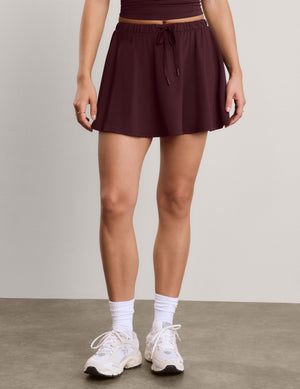 A model wearing a dark plum-colored athletic mini skirt with a drawstring waist, paired with white socks and white sneakers, standing against a neutral studio background.