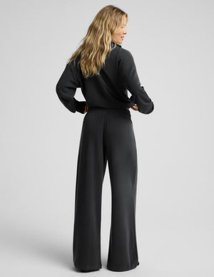 A woman with blonde hair is shown from the back wearing a black long-sleeve  top and matching wide-leg pants against a light gray studio background.