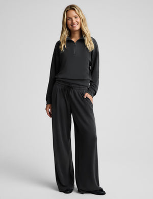 A woman modeling a matching dark gray lounge set consisting of a quarter-zip long-sleeve top and wide-leg sweatpants, photographed against a solid light gray studio background.