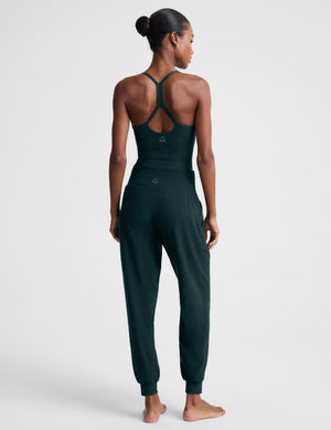 A woman modeling a dark forest green two-piece loungewear set consisting of a cropped tank top and matching jogger pants, shot against a light gray studio background.