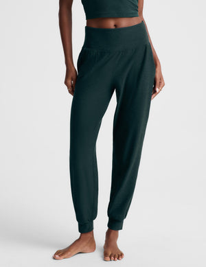 A woman modeling a dark forest green two-piece loungewear set consisting of a cropped tank top and matching jogger pants, shot against a light gray studio background.
