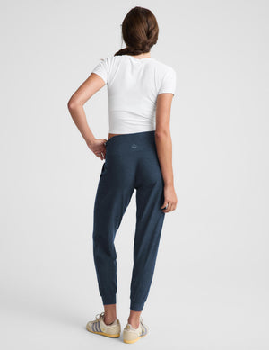 A woman modeling dark navy blue jogger pants with a white cropped t-shirt and beige sneakers, standing against a light gray studio background.