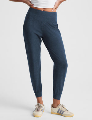 A woman modeling dark navy blue jogger pants with a white cropped t-shirt and beige sneakers, standing against a light gray studio background.
