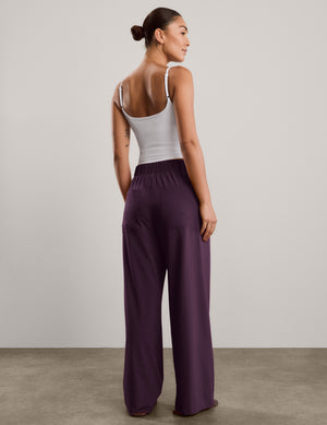 A woman shown from the back wearing a white tank top and dark purple wide-leg pants, standing against a neutral studio backdrop.