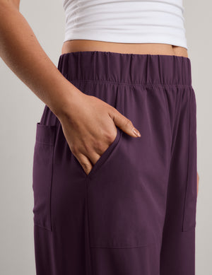 A woman shown from the back wearing a white tank top and dark purple wide-leg pants, standing against a neutral studio backdrop.