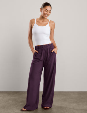 A woman shown from the back wearing a white tank top and dark purple wide-leg pants, standing against a neutral studio backdrop.