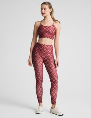 A woman modeling a matching red and pink plaid activewear set, consisting of a sports bra and high-waisted leggings, paired with white sneakers, against a light gray studio background.