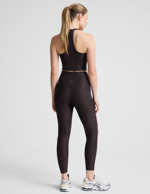 A woman modeling a matching athletic set consisting of a dark brown leopard crop tank and high-waisted leggings, paired with white and gray running shoes, against a light gray studio background.